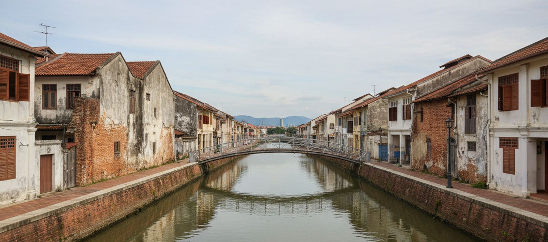 Wide view along the Melaka river between brick embankment walls and the backs of low shophouses, a footbridge crossing further upstream