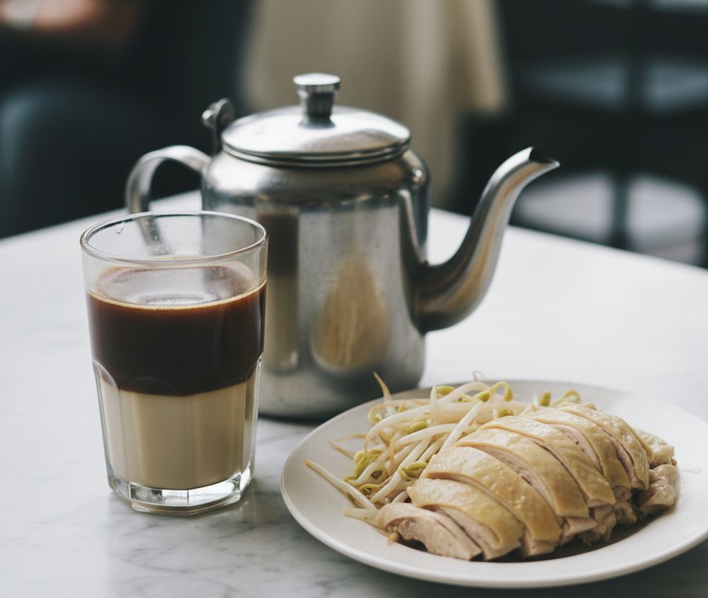 A marble topped table with a thick glass of dark coffee over condensed milk, a metal kettle, and a plate of poached chicken with blanched bean sprouts.