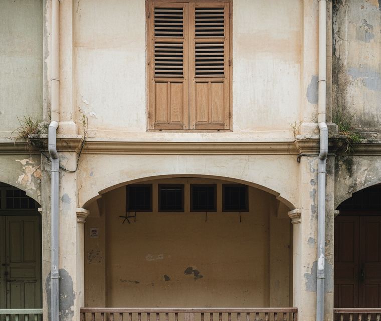 Weathered plaster facade: one closed louvered shutter upstairs, and an arch framing a shallow recess, four dark windows high in its back wall, a wooden balustrade across the base.
